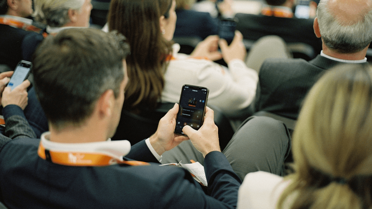 A seated audience wearing orange lanyards, reading a message on their phones